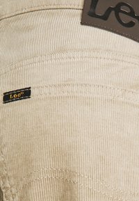 Béžová manšestrová látka s vertikálními pruhy, prošitým detailem a hnědou koženou nášivkou s názvem značky "Lee" v černé barvě.