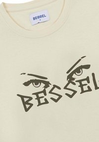 Camiseta de manga corta color crema con un diseño gráfico que presenta el texto en negrita "BESSEL" y ojos estilizados. Material de algodón, estilo cuello redondo.