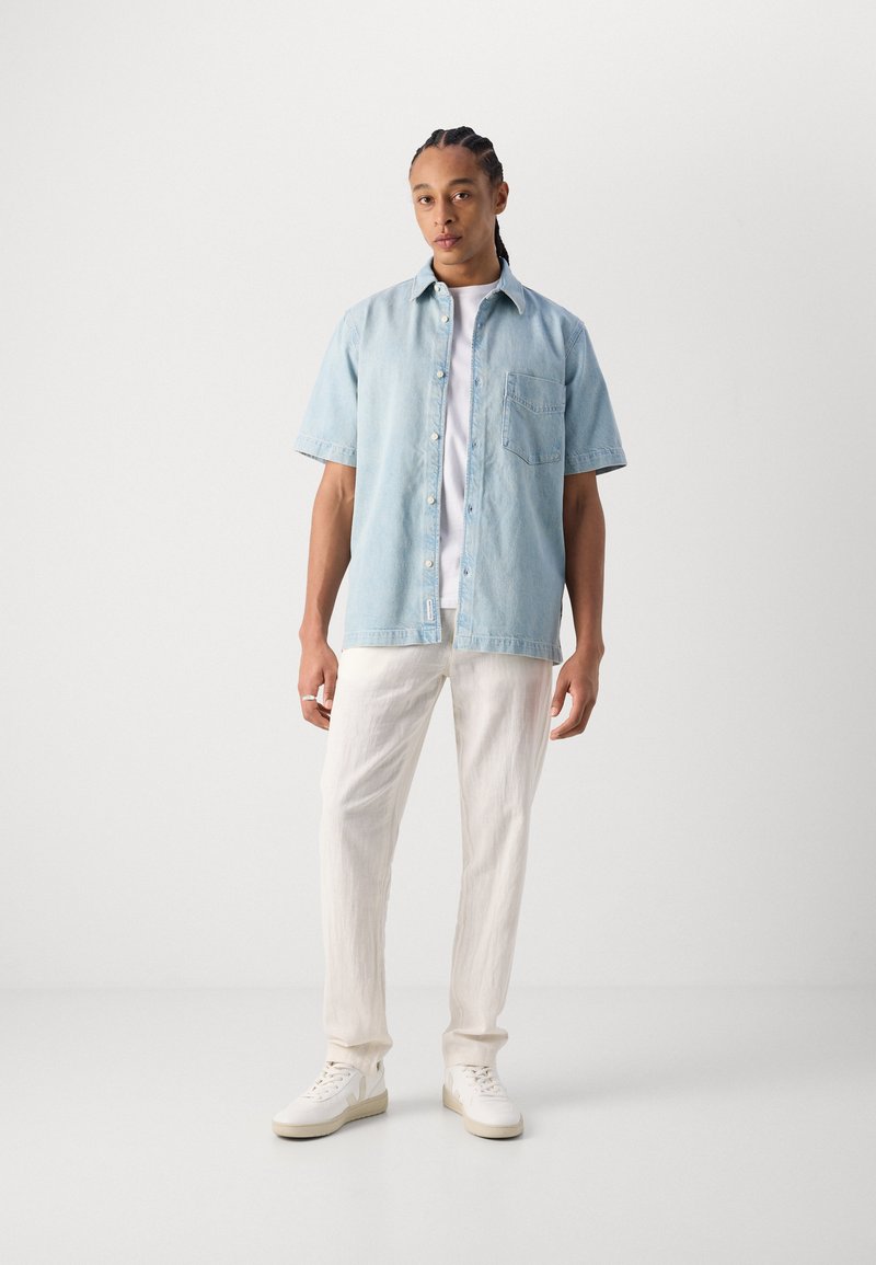 Camisa de mezclilla azul claro de manga corta con un bolsillo en el pecho, llevada sobre una camiseta blanca. Combinada con pantalones blancos texturizados y zapatillas.