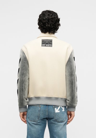 Άτομο που φοράει κρεμ ζακέτα βαρσαρίας με γκρι μανίκια και μαύρες ρίγες, συνδυασμένη με μπλε τζιν παντελόνι που έχει λευκό λογότυπο βέλους στην πίσω τσέπη.