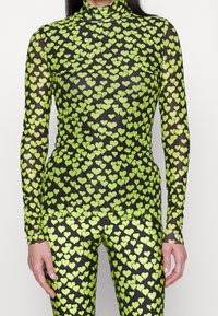 Langarm, durchsichtiger Mesh-Oberteil und passende Leggings mit einem grünen Herzmuster auf schwarzem Hintergrund. Verfügt über einen hohen Kragen und eine enganliegende Passform.