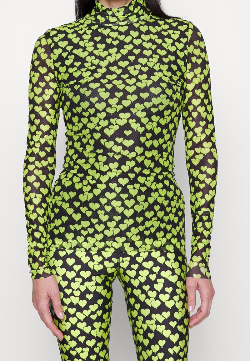 Langarm, durchsichtiger Mesh-Oberteil und passende Leggings mit einem grünen Herzmuster auf schwarzem Hintergrund. Verfügt über einen hohen Kragen und eine enganliegende Passform.