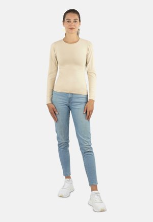 Junge Frau steht, trägt ein beiges Langarmoberteil, hellblaue Jeans, weiße Turnschuhe und goldene blattförmige Ohrringe vor einem schlichten Hintergrund.
