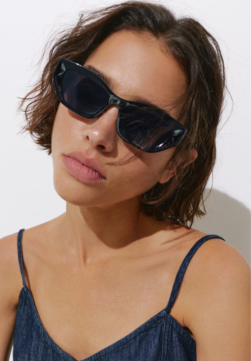 Femme aux cheveux courts bruns portant de grandes lunettes de soleil foncées et un top bleu à fines bretelles sur fond blanc.
