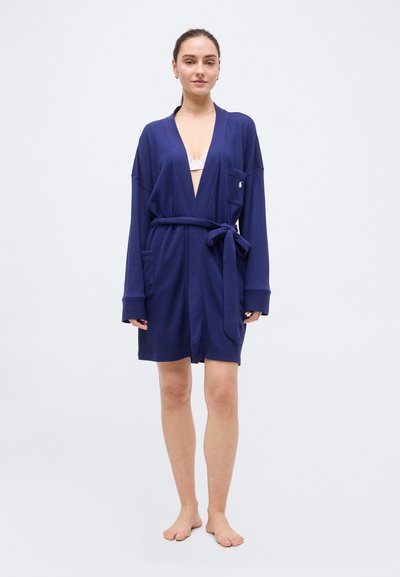 Γυναίκα όρθια, ξυπόλητη, φορώντας μια μπλε ναυτική ρόμπα με ζώνη πάνω από λευκή εσώρουχα, μπροστά σε απλό ανοιχτό φόντο.