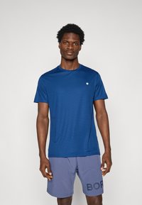 Navyblauw sportshirt met korte mouwen van ademend materiaal, met een klein logo op de borst, gecombineerd met lichtgrijze shorts met print.