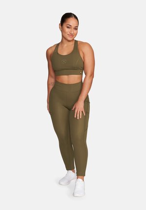 Kobieta stojąca i uśmiechająca się, ubrana w oliwkowozielony sportowy biustonosz i dopasowane legginsy z wysokim stanem oraz białe trampki na białym tle.
