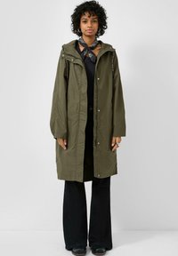 Vrouw met krullend haar die een olijfgroene capuchonjas draagt, een zwarte bandana-sjaal, een zwart shirt, zwarte broek met wijde pijpen en zwarte schoenen, staand tegen een witte achtergrond.