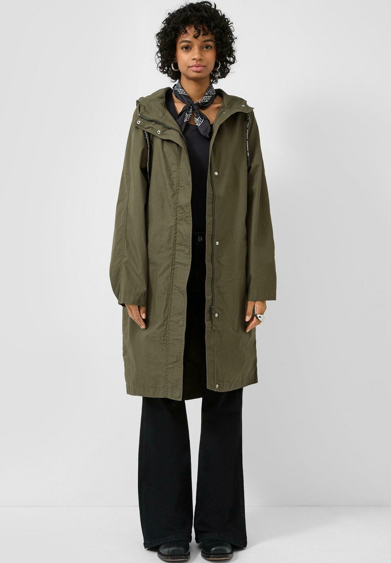 Vrouw met krullend haar die een olijfgroene capuchonjas draagt, een zwarte bandana-sjaal, een zwart shirt, zwarte broek met wijde pijpen en zwarte schoenen, staand tegen een witte achtergrond.