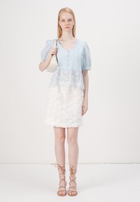 Blusa azzurro chiaro a maniche corte con dettagli in pizzo e gonna bianca a trama floreale. La modella tiene in mano una piccola borsa color crema, indossando sandali beige con lacci.