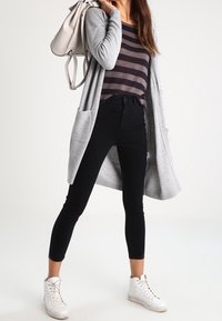Grauer Cardigan, gestreiftes Oberteil in Schwarz und Dunkelgrau, schwarze Slim-Jeans und weiße Sneaker. Der Cardigan hat Taschen und ein langes, fließendes Design.