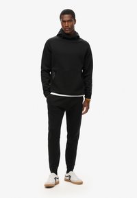 Sudadera negra con un bolsillo tipo canguro, puños elásticos y capucha con cordón, combinada con pantalones de chándal negros y zapatillas blancas con detalles en negro.