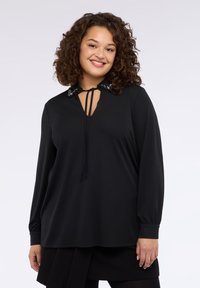 Czarna bluzka z długim rękawem i dekoltem w serek, ozdobiona dekoracyjnym kołnierzem z koralikami oraz wiązaniem przy szyi. Miękki materiał.