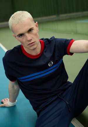 Maglietta blu navy con colletto rosso e righe blu, con logo sul lato sinistro, abbinata a pantaloni blu scuro e polsino, su un campo da tennis.