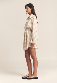 Beige-weiß kariertes Kleid mit elastischer Taille und Kragen, ausgestattet mit langen Ärmeln. Das Model trägt goldfarbene Flip-Flops. Einfaches Design.