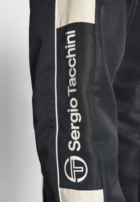 Pantalones deportivos negros con un acabado texturizado, que presentan un logo blanco en el costado y un acento en color crema en la parte superior.