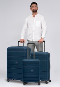 Uomo in camicia bianca e pantaloni grigi che tiene le maniglie di tre valigie rigide blu navy con ruote su sfondo neutro.
