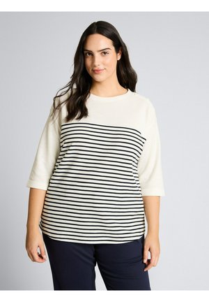 Kobieta z długimi ciemnymi włosami, ubrana w białą koszulę w czarne poziome paski oraz granatowe spodnie, stojąca na jednolitym tle.