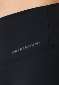 Black leggings made of stretchy fabric with a smooth texture. Features a logo in gray, positioned on the waistband.