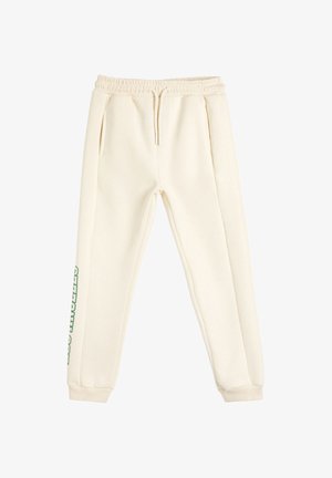 Pantalones jogger color blanco roto con cintura elástica, cordón, bolsillos laterales, puños acanalados y texto verde en la pierna izquierda.