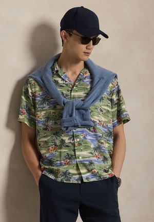 Koszula z krótkim rękawem w zielony kwiatowy wzór, z różowymi kwiatami i palmami. Niebieski sweter zarzucony na ramiona, w połączeniu z ciemnymi spodniami.