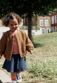 Brun, tekstureret fleecetrøje, rød- og hvidstribet skjorte, denimnederdel, farverige sneakers med blomsterdetaljer; udendørs omgivelser med græs.