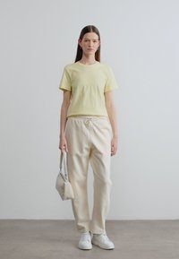 Ανοιχτό κίτρινο κοντομάνικο μπλουζάκι, συνδυασμένο με κρεμ φόρμες που διαθέτουν κορδόνι. Ένα μικρό λευκό τσαντάκι κρατιέται στο ένα χέρι.