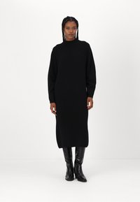 Abito nero lavorato a maglia con collo alto, maniche lunghe e silhouette dritta, che arriva a metà polpaccio. Indossato con stivali neri lucidi al ginocchio.