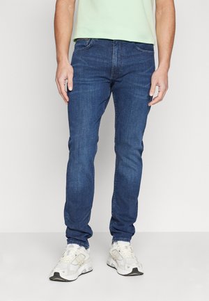 Jeans de mezclilla azul para hombre con un ajuste slim. Cuenta con cinco bolsillos, lazos para cinturón y una textura suave. Combinados con zapatillas deportivas blancas.