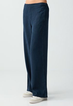 Persona con pantaloni blu scuro ampi abbinati a scarpe da ginnastica bianche, in piedi su un pavimento grigio chiaro contro uno sfondo semplice.