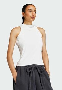 Biely bezrukávový športový top s vysokým golierom a jemným logom. Tkanina vyzerá hladko a priliehavo, čo dopĺňa voľné šedé tepláky.