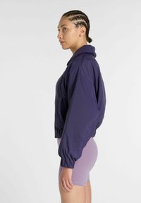Femme aux cheveux tressés portant une veste ample de couleur violet foncé et un short ajusté violet clair, debout de profil sur un fond blanc.
