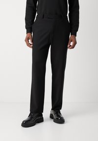 Pantalon noir ajusté avec fermeture à bouton et passants de ceinture, fabriqué à partir d'un tissu lisse. Porté avec des chaussures noires à lacets.