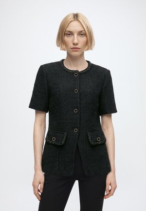 Frau mit kurzen blonden Haaren, die eine schwarze strukturierte Kurzarmjacke mit goldenen Knöpfen und zwei Vordertaschen trägt.
