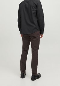 Mörk svart skjorta med knappar och långa ärmar, i kombination med mörkbruna byxor och svarta skor, sedd bakifrån. Slät yta, skräddarsydd passform.