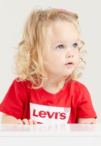 Rotes Baumwoll-T-Shirt mit einem weißen Logo und dem Text "Levi's". Das Kind hat lockige blonde Haare und die Hände ruhen auf einer weißen Oberfläche.