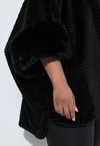 Poncho nero con cordini, dotato di morbidi polsini in pelliccia sintetica e caratterizzato da un motivo a zigzag testurizzato. Vestibilità ampia e comodo tessuto liscio.