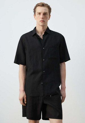 Młody mężczyzna stoi, ubrany w czarną koszulę z krótkim rękawem zapinaną na guziki oraz czarne krótkie spodenki na jednolitym jasnym tle.