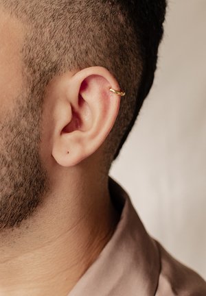 Orecchino a cerchio dorato sull'orecchio superiore, texture liscia, design circolare, indossato da una persona con capelli corti e castano chiaro e una barba ben curata.