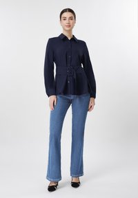 Γυναίκα που στέκεται, φοράει μπλε πουκάμισο μακρυμάνικο με ζώνη, μπλε τζιν bootcut και μαύρες μυτερές γόβες, μπροστά σε απλό λευκό φόντο.