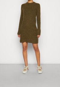 Olijfgroene gebreide jurk met lange mouwen, ronde hals en een lengte tot halverwege de dij. Gecombineerd met beige sneakers met een gestructureerd ontwerp.