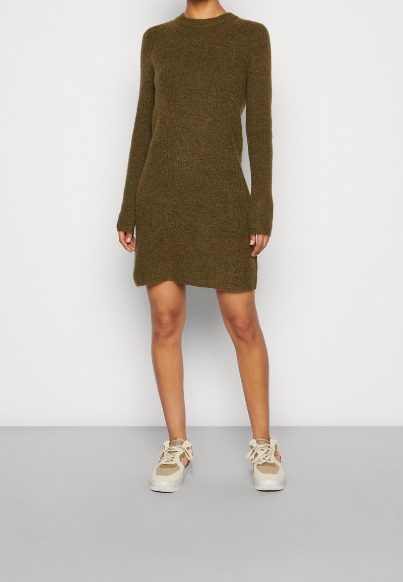 Olijfgroene gebreide jurk met lange mouwen, ronde hals en een lengte tot halverwege de dij. Gecombineerd met beige sneakers met een gestructureerd ontwerp.