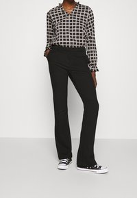 Schwarze Schlaghose kombiniert mit einer schwarz-weiß karierten Bluse mit V-Ausschnitt und Rüschen an den Manschetten. Getragen mit klassischen schwarzen Sneakern.