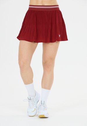 Kvinde iført et rødt plisseret skørt, hvide sportsstrømper og hvide sportssko står på en hvid baggrund.