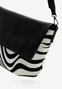 Musta nahkainen läppäkäsi, jossa on kohotettu brändin nimi ja mustavalkoinen abstrakti raidallinen kangasrunko. Tässä laukussa on irrotettava olkaintasku.