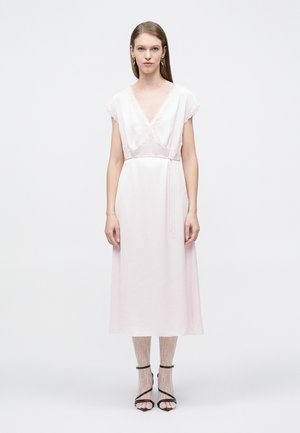 Γυναίκα με ανοιχτό ροζ μίντι φόρεμα με δαντέλα, καλσόν με δαντέλα και μαύρα πέδιλα με λουράκια, στέκεται μπροστά σε απλό λευκό φόντο.