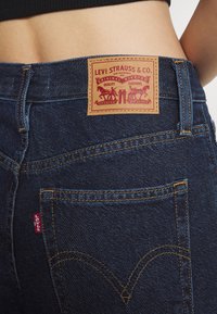 Vaqueros de denim azul oscuro con un diseño distintivo en el bolsillo trasero, una etiqueta de cuero marrón con gráficos rojos y detalles de costura amarilla.