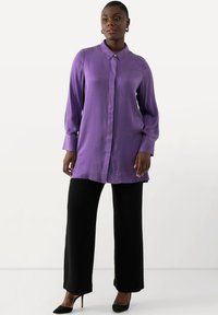 Blusa viola a maniche lunghe con colletto affilato, chiusura a bottoni e texture liscia, abbinata a pantaloni neri a gamba larga e tacchi.