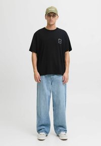 Jauns vīrietis ģērbts melnā pārspīlētā T-kreklā, gaiši zilās platas biksēs, baltās kedās un olīvzaļā cepurē, stāv pret viengabalainu fona.