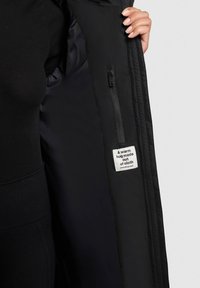 Černá péřová bunda s prošívanou texturou, zobrazující zipovou vnitřní kapsu a textilní štítek s nápisem "Teplé objetí vyrobené z látky."